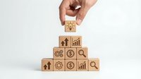 Hand placing wooden cube with lightbulb icon on pyramid of business blocks featuring growth arrows, gears, dollar signs and charts on white background.