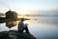 Man reading book by misty lake at sunrise with rustic cabin and chimney smoke creating peaceful morning atmosphere for relaxation and solitude concepts.