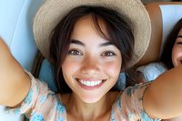 Young Asian woman wearing straw hat and floral dress taking selfie with bright genuine smile, close-up portrait showing natural beauty and joy.