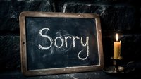 Chalkboard with the word Sorry beside a lit candle on a dark brick wall background, conveying apology, regret, and sincere emotion.