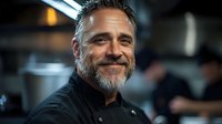 Smiling middle-aged Caucasian male chef with gray beard in black uniform standing confidently in professional restaurant kitchen during service.