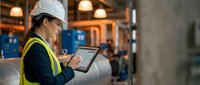 Female engineer in hard hat and safety vest using digital tablet at industrial construction site for project management and quality control.