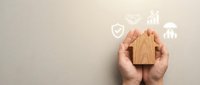Hands holding wooden house with security and financial growth icons floating above, representing home protection, insurance, investment, and real estate business concepts.