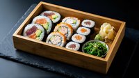 Assorted sushi rolls with salmon, tuna and avocado served in a wooden tray with seaweed salad and pickled ginger on dark slate.