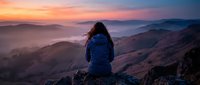 Woman hiker with backpack sitting on mountain peak at sunrise, overlooking misty valleys and distant hills. Adventure travel and outdoor exploration.