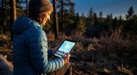 Young man using tablet computer outdoors in forest during golden hour sunset. Digital nomad working remotely in nature wilderness camping adventure.
