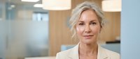 Professional mature Caucasian businesswoman with short blonde hair in beige blazer smiling confidently in modern office environment for corporate presentations.