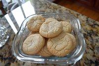 Fresh baked sugar cookies with crackled tops served on clear glass plate against granite counter, showcasing homestyle baking and comfort food appeal.