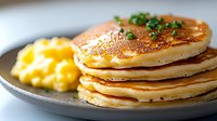 Stack of fluffy golden pancakes garnished with fresh herbs served on gray ceramic plate with side of scrambled eggs, close up view of breakfast food.