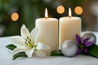 Glowing white candles with white lily and purple orchid flowers, silver Christmas ornaments on white tablecloth against soft bokeh background.
