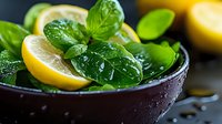 Fresh lemon slices and wet basil leaves with water drops in dark ceramic bowl against black background, moody food photography composition.