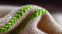 Textured natural linen fabric with decorative green woven stripes in close-up macro shot showing detailed textile structure and weaving pattern against blurred background.
