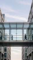 Modern glass skybridge connecting office buildings with silhouettes of business professionals walking across transparent walkway in urban setting.