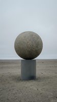 Large granite sphere balanced on concrete cylindrical pedestal in barren coastal landscape with sandy beach and overcast sky, suitable for contemporary art, architectural visualization, and minimalist design projects.