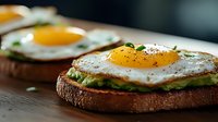 Sunny side up eggs on toasted bread with mashed avocado, seasoned with black pepper and fresh herbs, served on rustic wooden surface in moody lighting.
