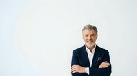 Mature male professional with gray hair and light complexion wearing dark navy blazer over white shirt, arms crossed, smiling confidently against white background. Corporate portrait for business, leadership, executive profiles.