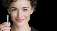 Young Caucasian woman with natural makeup and curly hair smiling while holding silver metallic lipstick against black background, beauty and cosmetics concept.