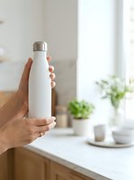 Person with medium skin tone holding white insulated water bottle with stainless steel cap in bright kitchen with plants and natural light for beverage, lifestyle and product marketing.