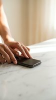 Woman's hands with rings reaching for a smartphone on a marble surface in a bright, cozy interior setting.