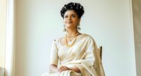 Woman with dark curly hair and medium skin tone wearing cream silk robe and gold jewelry, seated against neutral background for portrait, fashion, lifestyle, and beauty content.