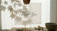 Hand holding a thank you card near a sunlit window with maple leaf shadows, autumn leaves and a ceramic bowl on a wooden table.