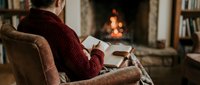 Woman in cozy red sweater reading book by warm fireplace in comfortable armchair creating peaceful winter evening atmosphere at home.