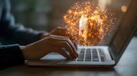 Person typing on laptop with magical glowing light bulb emerging from screen, surrounded by golden sparkles and energy particles in dark room setting.