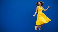 Young woman in bright yellow dress jumping with joy against blue background, expressing happiness and freedom in motion.