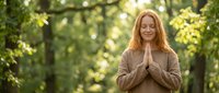 Middle-aged Caucasian woman with red hair practicing meditation in prayer pose outdoors in peaceful green park setting with natural sunlight filtering through trees.