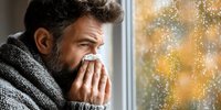 Middle-aged Caucasian man with beard blowing nose into tissue while looking through rainy window with autumn colors in background, depicting seasonal illness.
