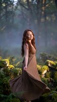 Young Caucasian woman with long brown hair in flowing beige dress standing peacefully in enchanted forest with sunbeams and moss-covered rocks for wellness content.