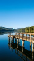 Wooden dock extending into calm alpine lake surrounded by forested mountains under clear blue sky, perfect for travel and nature backgrounds.