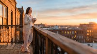 Young woman in casual white clothing standing on urban rooftop balcony at golden hour sunset, holding coffee cup, overlooking city skyline with warm evening light and buildings below.