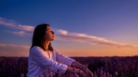 Woman with long dark hair, wearing a white shirt, sitting in a lavender field during golden hour sunset. Peaceful outdoor portrait for wellness, nature, and lifestyle content.
