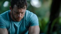 Muscular Caucasian man in turquoise t-shirt looking down thoughtfully against blurred green background, showing emotional vulnerability and contemplation in fitness setting.