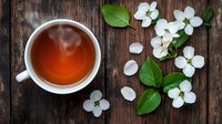 Steaming cup of aromatic tea with fresh white spring blossoms and green leaves scattered on rustic wooden background, overhead view for food styling.