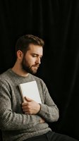 Thoughtful young man in knit sweater holding white book against dark background, contemplating reading in warm natural light for education concepts.