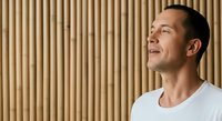 Middle-aged man with dark hair and facial stubble wearing white casual t-shirt, smiling while looking upward against beige corrugated wall background, expressing optimism and positivity for lifestyle and motivational content.