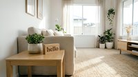Bright minimalist living room with beige sofa, wooden coffee table, hello sign, and green plants by a sunlit window in a cozy home interior.