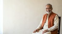 Senior man with gray beard and glasses wearing orange vest over white shirt, seated against neutral background, suitable for corporate, business, professional portraits, and lifestyle content.