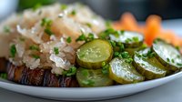 Grilled meat topped with sauerkraut, served with pickled cucumbers and fresh herbs on white plate, close up view of traditional European cuisine.