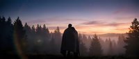 Silhouetted figure in dark cloak standing in misty forest at dawn, surrounded by evergreen trees and golden sunrise light breaking through fog and atmosphere.
