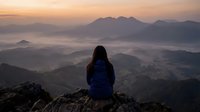 Woman with long dark hair wearing blue jacket sitting on rocky mountain peak overlooking misty valley and distant mountains at sunrise or sunset for travel, adventure, and nature content.