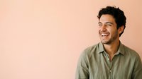 Young man with dark hair and olive complexion wearing sage green button-up shirt, smiling against peachy background. Suitable for lifestyle, business, and portrait projects.