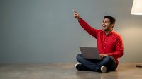 Young man with dark hair and South Asian ethnicity wearing red shirt sitting on floor with laptop, pointing upward in gesture of idea or success, indoor office setting with warm lighting.