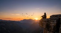 Person sitting on rocky cliff edge watching sunset over mountain landscape with flying birds in golden sky for meditation and adventure concepts.