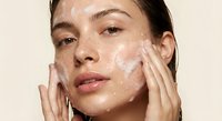 Young woman applying white facial cleanser cream with hands during skincare routine against neutral background for beauty and wellness concepts.