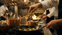 Professional chef pouring olive oil into sizzling pan while cooking in busy restaurant kitchen. Culinary expertise and gourmet food preparation in action.