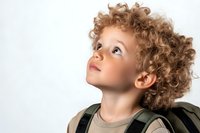 Young Caucasian child with curly blonde hair and expressive eyes looking up with wonder and curiosity against white background, wearing casual attire with backpack straps visible.