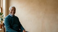 Senior African American man with gray hair and beard wearing dark teal sweater, sitting indoors near window with potted plant, smiling at camera. Portrait for lifestyle, wellness, retirement, and senior living concepts.